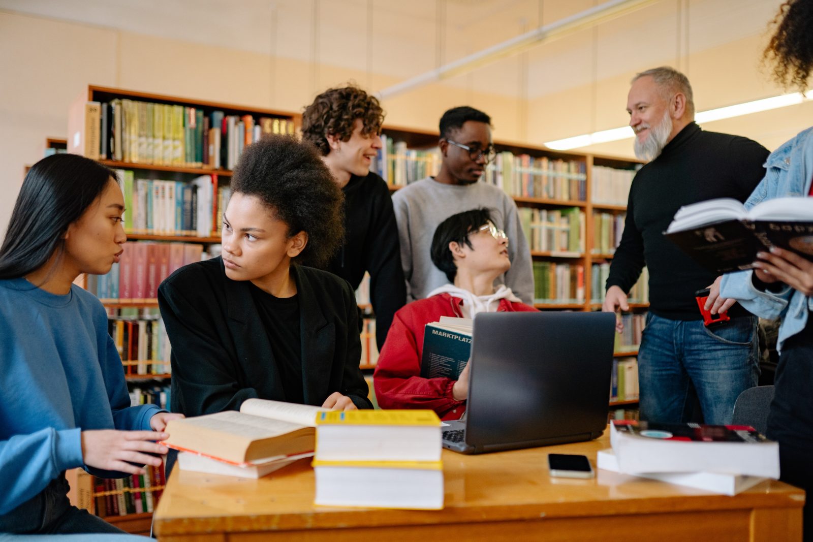 Diverse group of students crowded around a table in a library with a teacher
